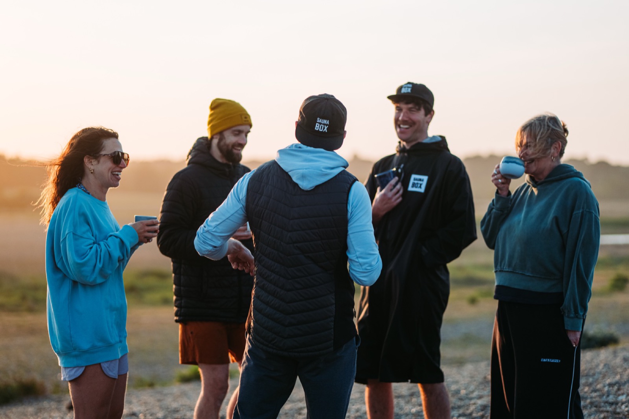 A group of friends in Sauna Box branded clothing laughing together at sunset, holding warm drinks after a sauna session
