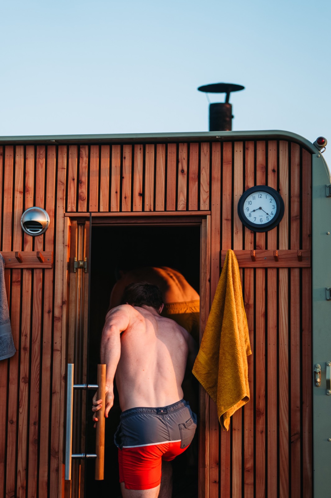 A man stepping into a cedar-clad wood-fired sauna in warm evening light, towel and clock beside the door