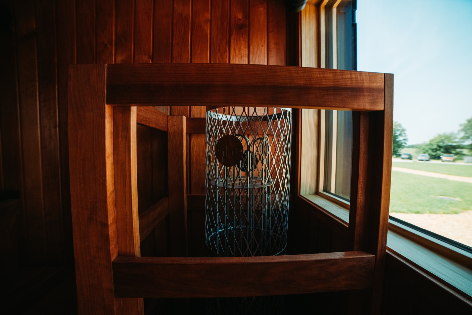 IKI stone heater in its mesh guard beside a window inside a Sauna Box container sauna, green field beyond